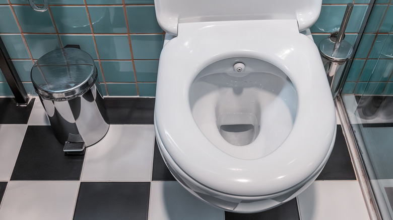 Clean white toilet bowl with built in bidet nozzle, metal trash can, and checked floor tiles in a modern lavatory