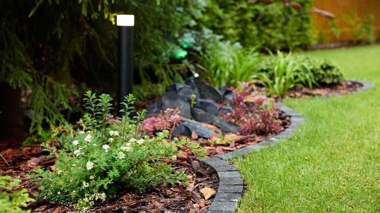A nicely landscaped garden with plants surrounded by mulch and small dark gray pavers separating it from a lawn