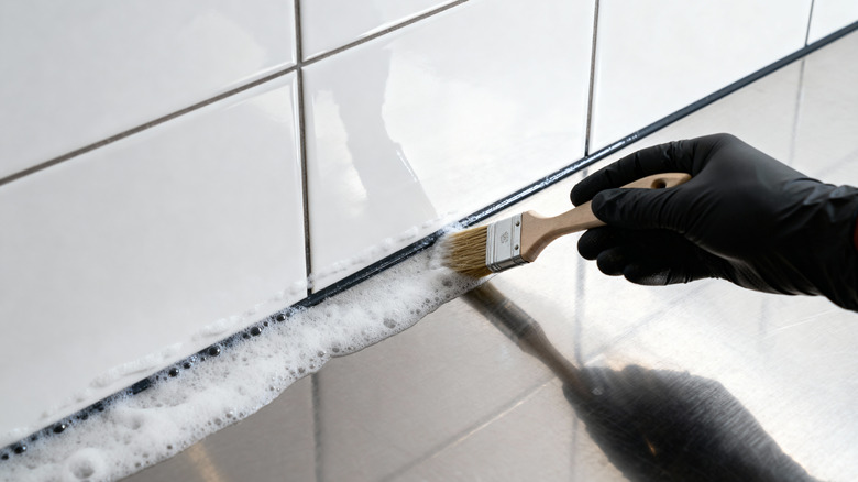 A gloved hand applying a foamy cleaning solution to tile grout