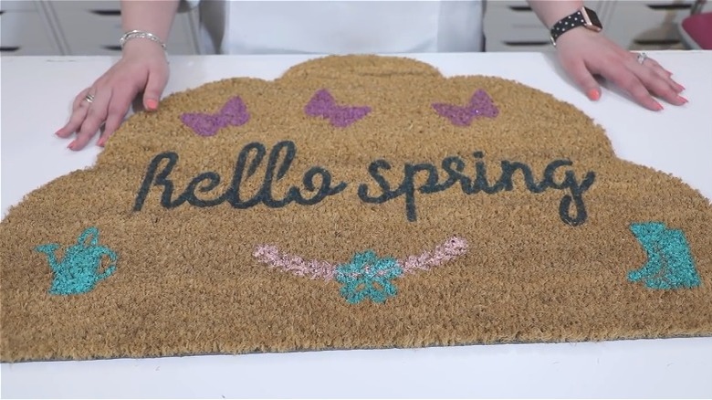 Woman's hands resting on table beside coir door mat she has painted with spring designs.