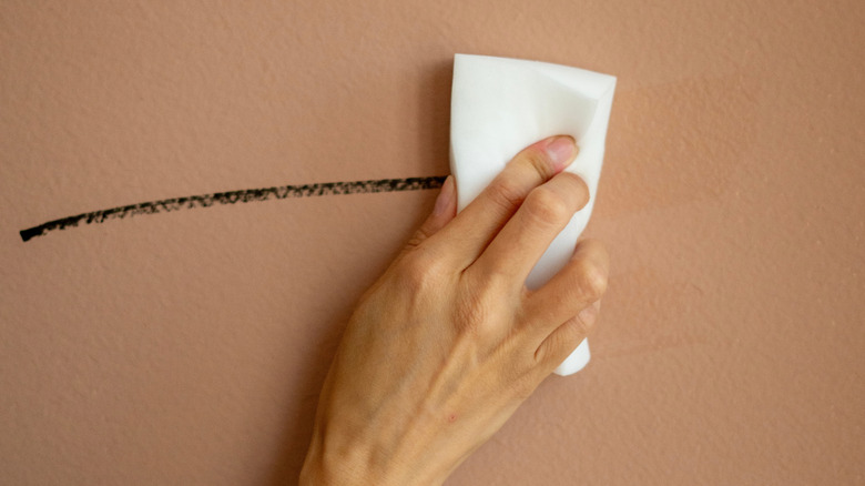 A close-up photo shows a hand cleaning a mark off a painted wall with a melamine foam sponge