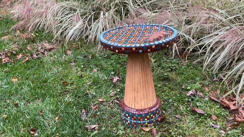 Birdbath covered in mosaic tiles sitting in on the ground with decorative grasses in the background