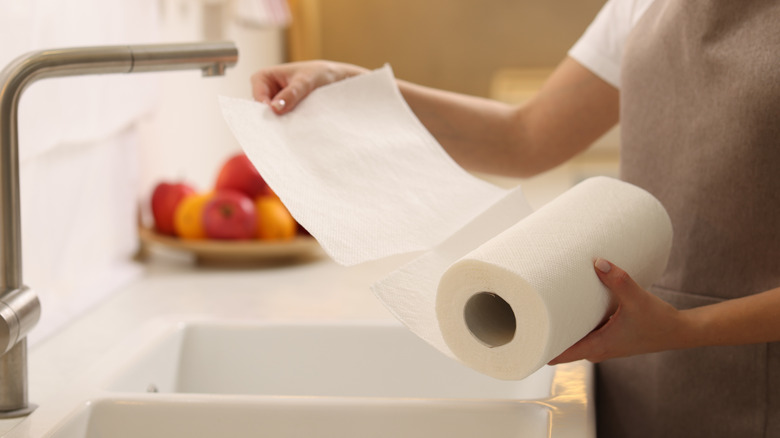 A person pulls a paper towel from a roll while standing next to the kitchen sink.