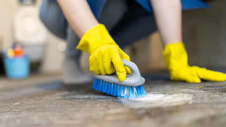 A person kneels on the floor, using a brush to scrub the surface while wearing yellow rubber gloves