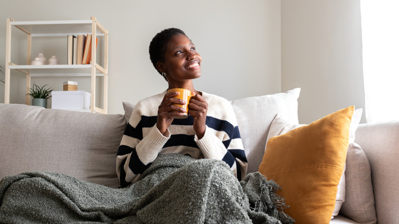 A woman sits on a gray sofa holding a mug and covered in a gray blanket as she looks towards the window
