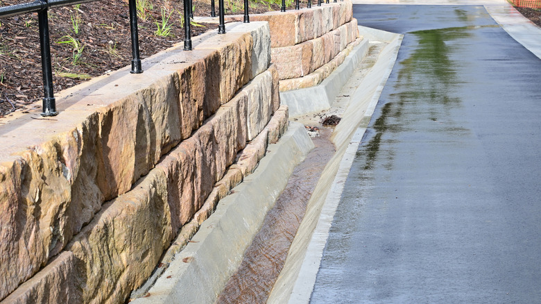 A sandstone retaining wall stands next to a drain during rainy weather