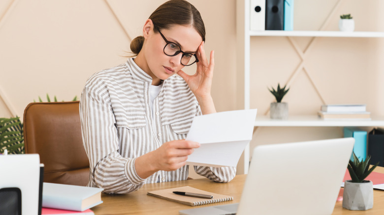 Concerned woman wearing glasses sitting in front of laptop staring at document she's holding