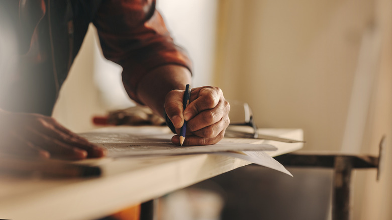 Hands writing on a paper with a penscil over raw wood planks over a sawhorse in a room