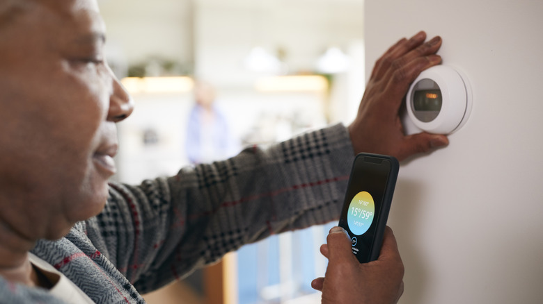 Person standing in front of smart thermostat and using a smartphone to adjust the settings