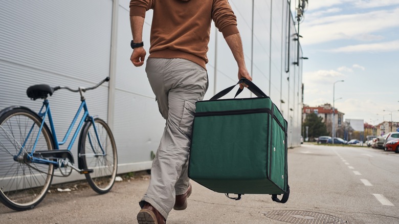 A delivery worker walks along a city street carrying a large green insulated bag, with a bicycle parked nearby